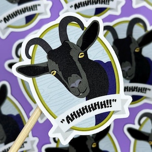 May include: A sticker featuring a black goat head with curved horns, yellow eyes, and a blue collar. The goat is inside a gold-lined oval with a light blue background. The sticker has the text "AHHHHHH!!" on a white banner.