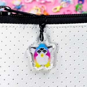 May include: A clear acrylic charm featuring a colorful Furby design with a black clasp. The charm has a white body with a rainbow gradient and a blue, pink, and yellow color scheme. The charm is attached to a black zipper.