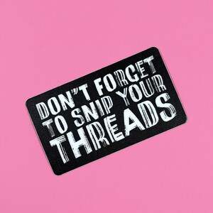 May include: A black rectangular sticker with rounded corners, featuring white text that reads "DON'T FORGET TO SNIP YOUR THREADS" against a black background. The sticker is set against a bright pink background.