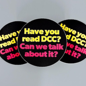 Könnte beinhalten: Drei kreisförmige schwarze Aufkleber mit gelbem und rosa Text. Der Text lautet: Have you read DCC? Can we talk about it? Der gelbe Text befindet sich in den oberen zwei Zeilen und der rosa Text in den unteren zwei Zeilen, alles in Fettschrift.