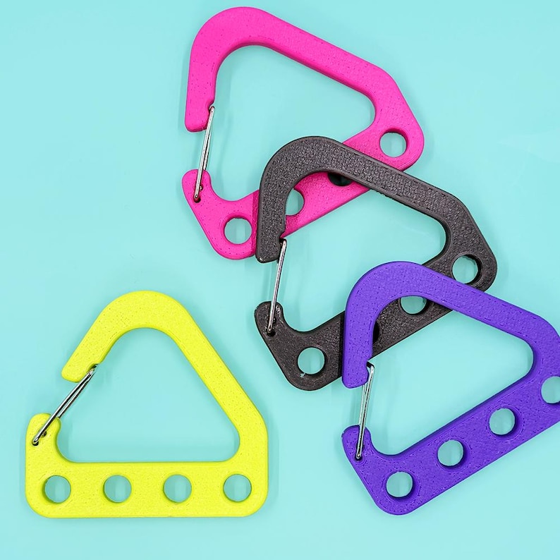 May include: Four colorful carabiner clips in pink, black, purple, and yellow. The clips have a triangular shape with a spring-loaded gate and multiple circular holes. The background is a solid light blue.