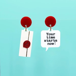May include: A pair of novelty earrings with red circular studs. One earring is a white rectangle with a red wax seal and gold string. The other is a white speech bubble with the text "Your time starts now!" in black.