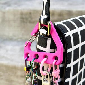 May include: A bright pink key holder with multiple key rings and clasps attached to a black and white patterned purse strap. The key holder is triangular with several holes for attaching keys and accessories.
