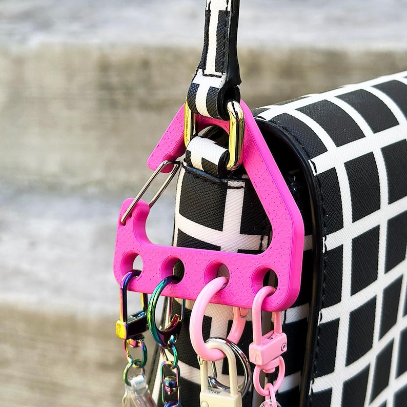 May include: A bright pink key holder with multiple key rings and clasps attached to a black and white patterned purse strap. The key holder is triangular with several holes for attaching keys and accessories.