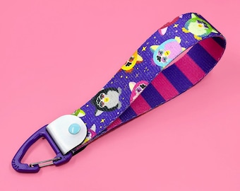 Puede incluir: Una correa de muñeca morada con un patrón de personajes de dibujos animados y un mosquetón morado. La correa presenta un patrón repetido de personajes de dibujos animados coloridos sobre un fondo morado oscuro con estrellas amarillas. El clip está sujeto a un botón blanco.