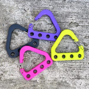 May include: A collection of colorful, triangular carabiner clips in black, purple, pink, and yellow. Each clip has a metal spring gate and four circular holes. The clips are arranged on a textured gray surface.