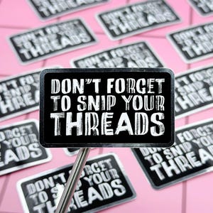 May include: A collection of black and white rectangular stickers with the text "DON'T FORGET TO SNIP YOUR THREADS". The stickers are on a pink background. One sticker is mounted on a silver stick.