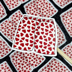 May include: A sticker featuring a pair of white boxer shorts covered in red hearts. The shorts have a black waistband and are set against a background of similar stickers.
