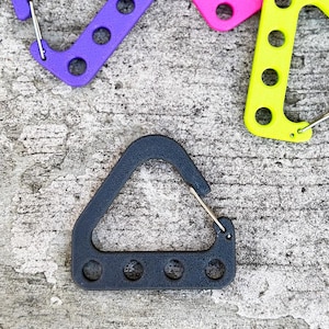May include: A dark gray carabiner with a triangular top and a rectangular bottom featuring four circular holes. The carabiner has a silver metal clasp. Other colorful carabiners in purple, pink, and yellow are visible in the background.