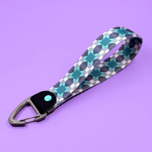 May include: A patterned wrist strap with a grey carabiner clip. The strap features a repeating geometric design in teal, grey, and white. The strap is attached to a black leather piece with a teal dot.