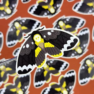 May include: A black and yellow moth sticker with a skull design. The moth has white accents on its wings and a yellow body. The sticker is on a red background.