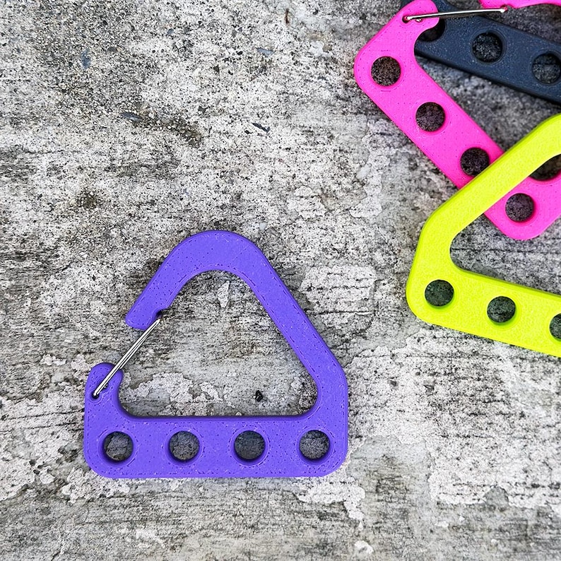 May include: A close-up shot of several colorful carabiner-style clips. The primary clip is purple, with a silver metal clasp. Other clips are pink, black, and yellow. Each clip has four circular holes. The clips are on a textured gray surface.