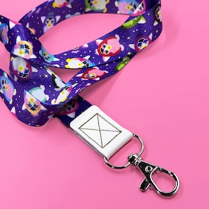 May include: A purple lanyard featuring a repeating pattern of colorful cartoon characters. The lanyard has a white leatherette tag with a stitched design and a silver-colored metal clasp.