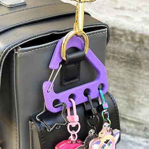 May include: A purple key holder with a triangular design is attached to a black bag with a gold-colored clasp. Several keychains with various charms are attached to the key holder, including a pink heart and a black star.