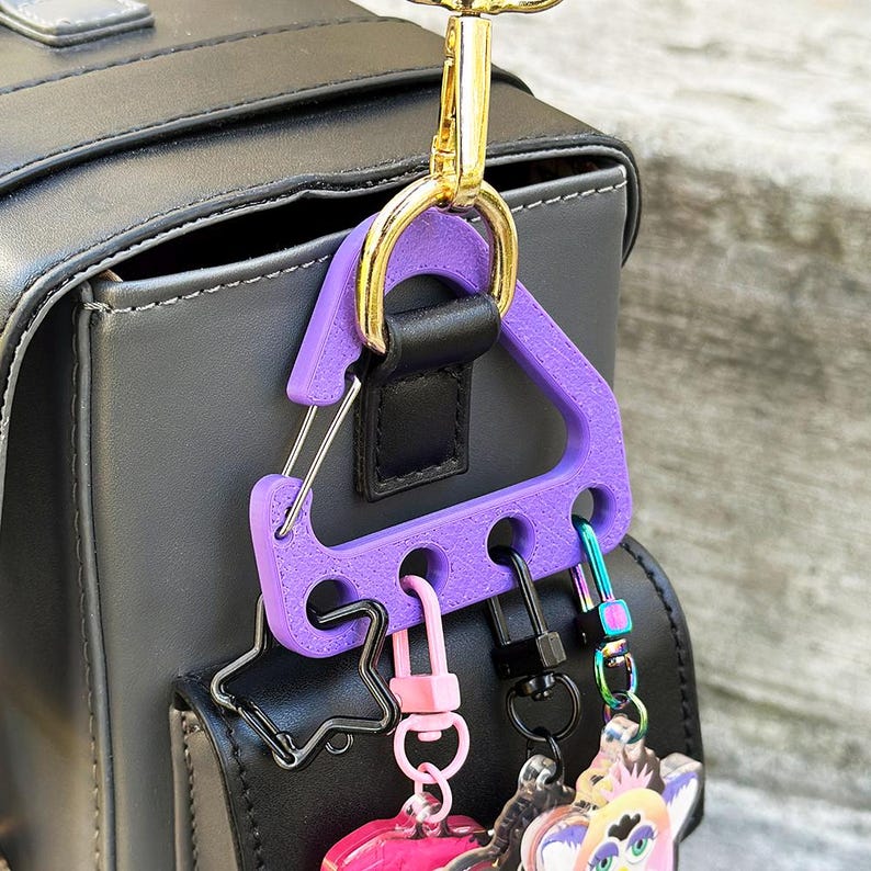 May include: A purple key holder with a triangular design is attached to a black bag with a gold-colored clasp. Several keychains with various charms are attached to the key holder, including a pink heart and a black star.