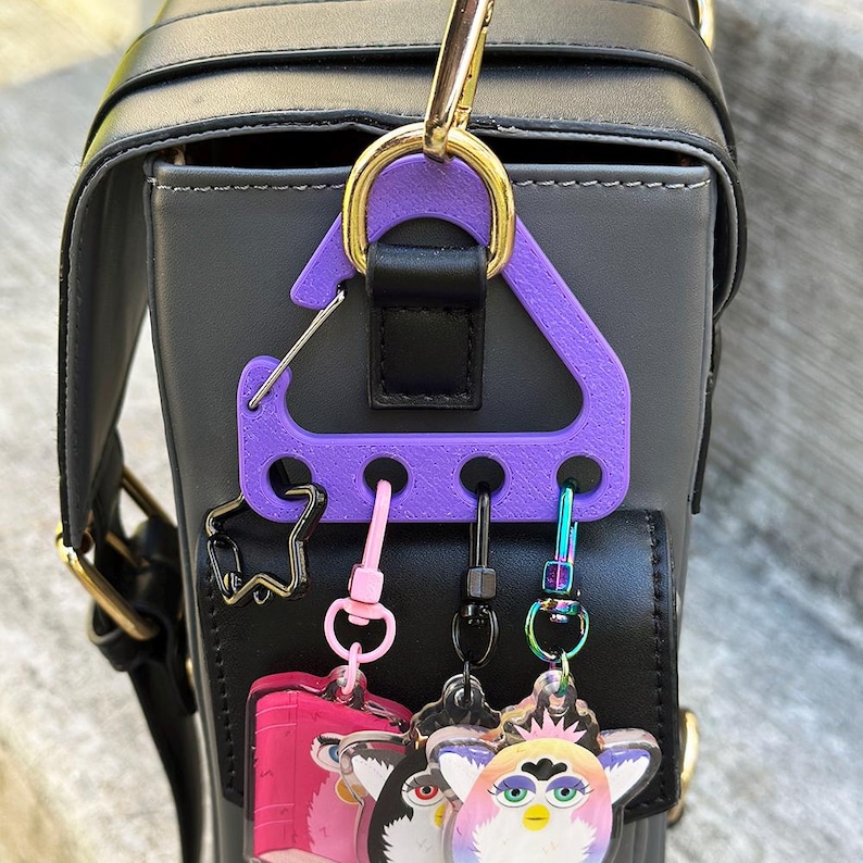 May include: A black and gray handbag with a gold-colored handle and a purple keychain holder. The keychain holder has several charms, including a pink bus and Furby-themed keychains. The bag has a black strap.