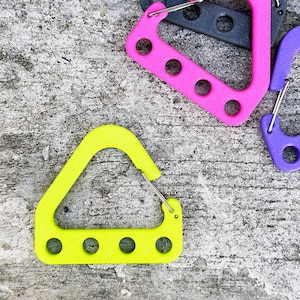 May include: A close-up shot of several colorful carabiner clips. The clips are in various colors, including neon yellow, pink, purple, and gray. Each clip has a metal spring-loaded gate and a series of circular holes. The clips are arranged on a textured gray surface.
