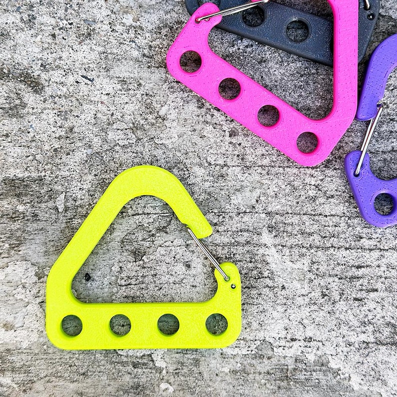 May include: A close-up shot of several colorful carabiner clips. The clips are in various colors, including neon yellow, pink, purple, and gray. Each clip has a metal spring-loaded gate and a series of circular holes. The clips are arranged on a textured gray surface.