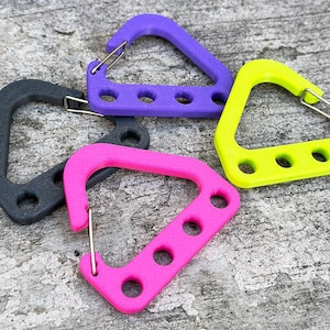 May include: A collection of colorful carabiner clips in black, purple, pink, and yellow. Each clip has a triangular shape with three circular holes and a metal spring gate. The clips are arranged on a textured gray surface.