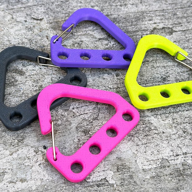 May include: A collection of colorful carabiner clips in black, purple, pink, and yellow. Each clip has a triangular shape with three circular holes and a metal spring gate. The clips are arranged on a textured gray surface.