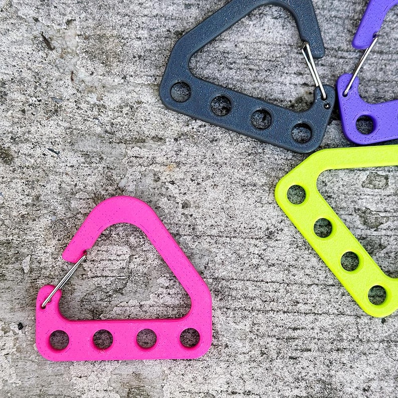 May include: A collection of colorful carabiner clips in various shades, including pink, gray, purple, and neon green. Each clip has a triangular or rectangular shape with multiple circular holes and a metal spring gate for secure closure. The clips are designed for attaching items.