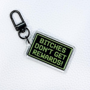 May include: A rectangular keychain with the text "BITCHES DON'T GET REWARDS!" in green on a black background. The keychain has a clear border and a black metal clasp.