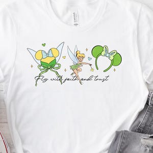 Puede incluir: Camiseta blanca con un gráfico de Campanilla, diademas de orejas de ratón y el texto "Fly with faith and trust". El diseño incluye detalles en amarillo, verde y azul. La camiseta se combina con vaqueros grises y zapatillas blancas.