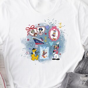 May include: A white t-shirt featuring a graphic print of Disney characters including Mickey Mouse, Minnie Mouse, Donald Duck, and Goofy. The design includes a cruise ship, nautical icons, and decorative frames against a blue watercolour splash background.