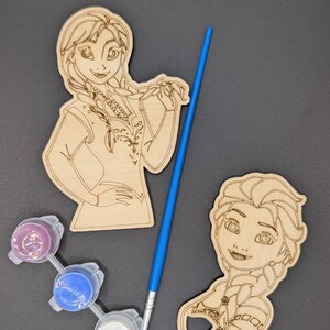 May include: Two laser-cut wooden character cutouts featuring engraved outlines of figures with braided hair, accompanied by a blue-handled paintbrush and a small plastic strip containing three circular paint pots in purple, blue, and white.