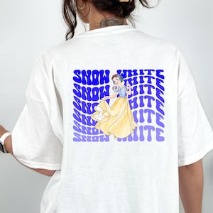 Puede incluir: Camiseta blanca con un estampado gráfico azul y blanco de Blancanieves. El gráfico presenta a Blancanieves con un vestido amarillo con un corpiño azul y un lazo rojo en el pelo. El texto "SNOW WHITE" se repite en una fuente azul ondulada.