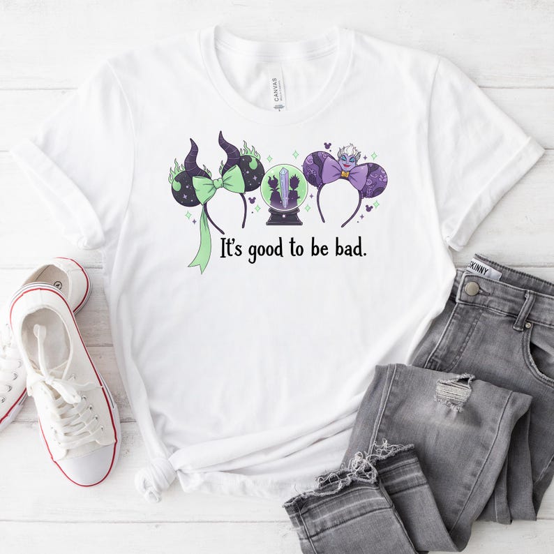 May include: White t-shirt featuring a graphic of three character-themed mouse ears with a crystal ball. The ears are green, purple, and black. The text "It's good to be bad" is printed below the graphic. A pair of white sneakers and gray jeans are also visible.