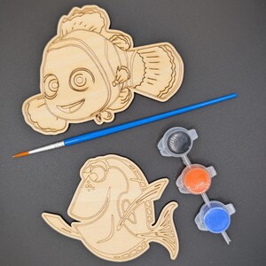 May include: Two laser-cut wooden fish shapes featuring engraved outlines of a clownfish and a tang fish. The set includes a thin paintbrush with a blue handle and orange bristles, along with three small connected plastic paint pots containing black, orange, and blue paint.