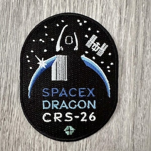 May include: Black oval patch featuring a SpaceX Dragon CRS-26 mission design. The patch includes a stylized rocket, the International Space Station, and the SpaceX logo. The design incorporates blue and white accents against a starry background.
