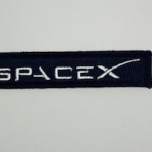 May include: A black rectangular patch featuring the word "SPACEX" embroidered in white, accompanied by a white stylized "X" symbol. The patch is probably made of fabric and has a stitched border.