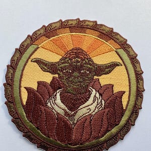 May include: Embroidered patch depicting a stylized Yoda character with three eyes, set within a lotus flower design. The patch is circular, with a brown, green, and yellow colour scheme. The background features a sunburst pattern.