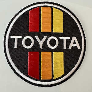 May include: A black and white embroidered patch with the word "TOYOTA" in white lettering and three vertical stripes in red, orange, and yellow.