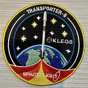 Könnte beinhalten: Ein gestickter Patch in Schwarzweiß mit einer Rakete, die von der Erde abhebt. Der Patch zeigt die Aufschrift "TRANSPORTER-6" und "KLEOS" mit einem roten und gelben "SPACEFLIGHT" am unteren Rand.
