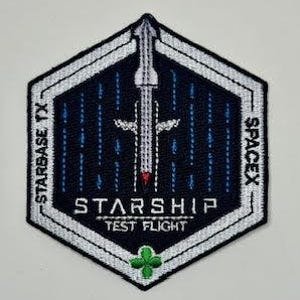 May include: A black and white embroidered patch with a white hexagonal frame. The patch features a white rocket ship with a red tip, surrounded by blue and white lines. The text "Starship Test Flight" is written in white below the rocket ship. The text "SpaceX" is written in white on the right side of the patch and "Starbase TX" is written in white on the left side of the patch.