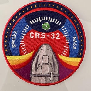 Könnte beinhalten: Runder, gestickter Aufnäher mit einem Raumschiffdesign und dem Text "CRS-32". Der Aufnäher hat ein rotes, blaues und gelbes Farbschema. Die Wörter "SPACEX" und "NASA" sind ebenfalls sichtbar.