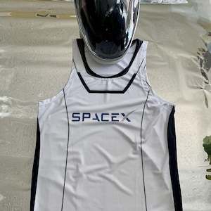 Könnte beinhalten: Ein weißes ärmelloses Shirt mit schwarzem Besatz und dem in Blau gedruckten Wort "SpaceX". Das Shirt hat außerdem eine kleine amerikanische Flagge und ein NASA-Logo.