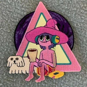 Op de afbeelding: Geborduurde patch met een stripfiguur met een grote roze hoed, die een kopje vasthoudt en een koptelefoon draagt. Links is een witte hond en een roze driehoek en een paarse cirkel vormen de achtergrond. De patch ligt op een grijze ondergrond.