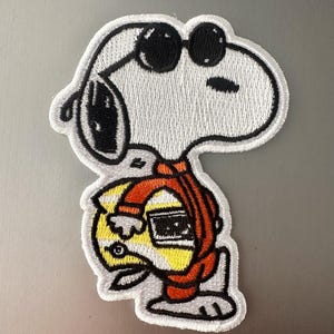 May include: Embroidered patch featuring Snoopy wearing sunglasses and a yellow and orange outfit. The patch has a white background with black, yellow, and orange details. The patch is likely for a jacket, backpack, or other fabric item.