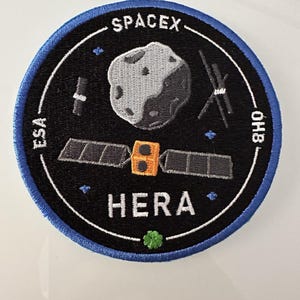 Könnte beinhalten: Ein gestickter schwarzer und weißer Aufnäher mit blauem Rand. Der Aufnäher zeigt ein Raumschiff, einen großen Asteroiden und den Text "SPACEX", "ESA", "OHB" und "HERA".
