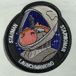 May include: A black and white embroidered patch with a cartoon astronaut wearing a helmet. The patch has the text "Starman Launchmankind Starwoman" around the edge.