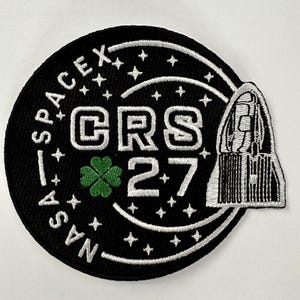 Peut inclure: Un écusson brodé noir et blanc avec le texte "NASA - SPACEX CRS 27" et un trèfle à quatre feuilles vert. L'écusson présente également un contour blanc d'une fusée.