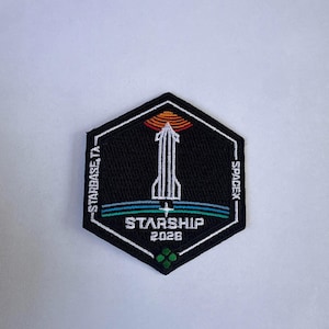 May include: A hexagonal embroidered patch with a black background featuring a white outline of a rocket ship. The design includes an orange sun, blue and green horizontal lines, and the text STARBASE, TX, SPACEX, STARSHIP, and 2026 in white.