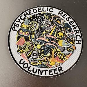Op de afbeelding: Geborduurde ronde patch met de tekst "PSYCHEDELIC RESEARCH VOLUNTEER". De patch toont kleurrijke illustraties van paddenstoelen, aliens en andere psychedelische beelden. Met een witte rand.