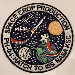 May include: Embroidered patch with a circular design featuring the text "SPACE CROP PRODUCTION" and "PH-04: HATCH TO ISS NASA KSC". The patch includes images of a rocket, Earth, a moon, and a flower, set against a starry background.