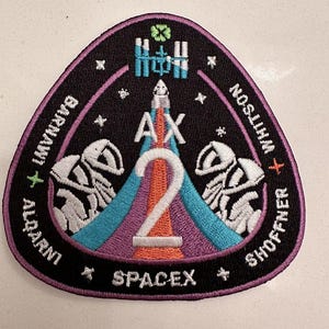Puede incluir: Un parche triangular negro y morado con el texto "SPACEX" y "AX-2" en blanco. El parche también presenta dos figuras blancas de astronautas, un trébol de cuatro hojas verde y los nombres de los astronautas: "LARRY CONNOR", "EYTAN STIVY", "HAYATO  YAMAZAKI" y "WALTER  WILSON".