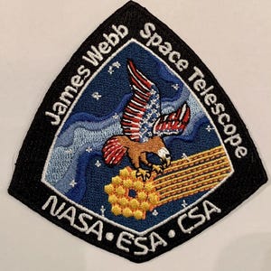 Puede incluir: Un parche bordado en blanco y negro con un águila azul, blanca y roja volando sobre una estructura de panal de abeja dorada. El texto "James Webb Space Telescope" está escrito en un arco blanco alrededor del águila. El texto "NASA - ESA - CSA" está escrito en un arco blanco en la parte inferior del parche.